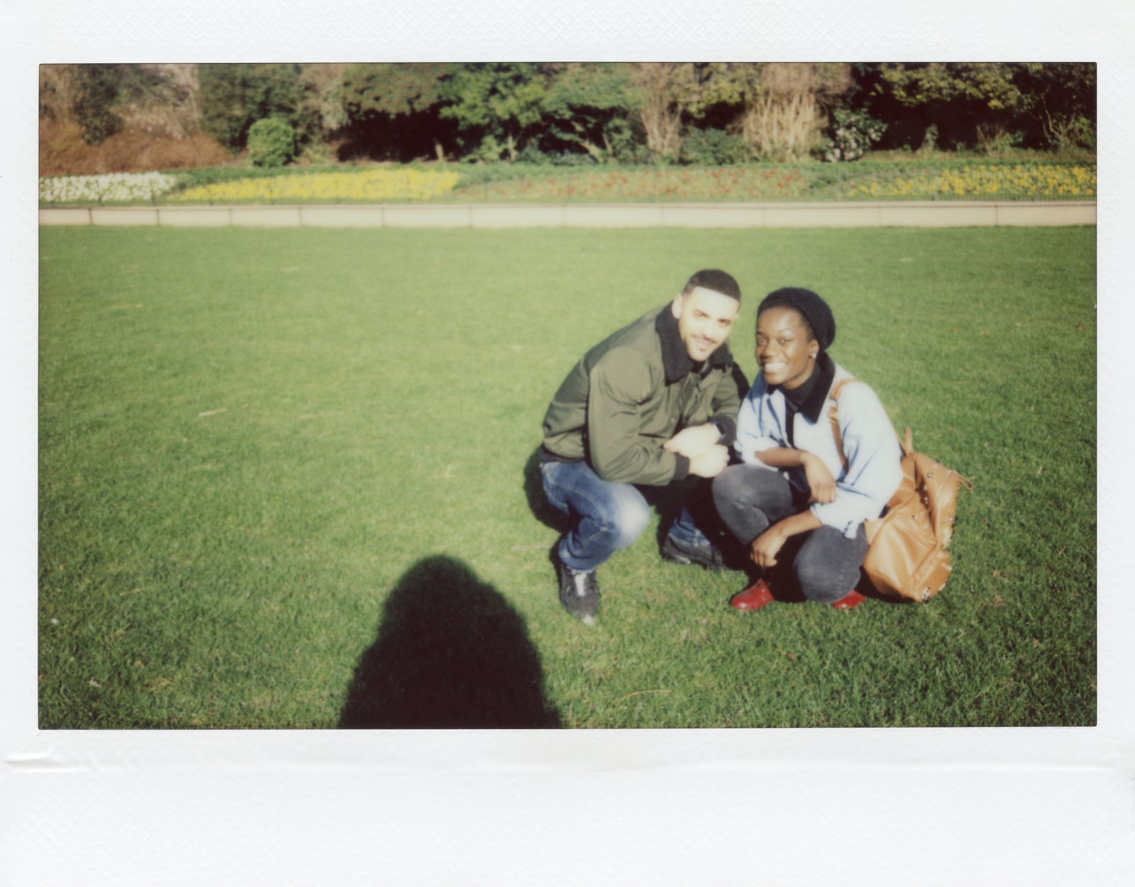 A polaroid-style grainy photo with a man and woman wearing denim pants and jackets sitting in a grassy park smiling at the camera.