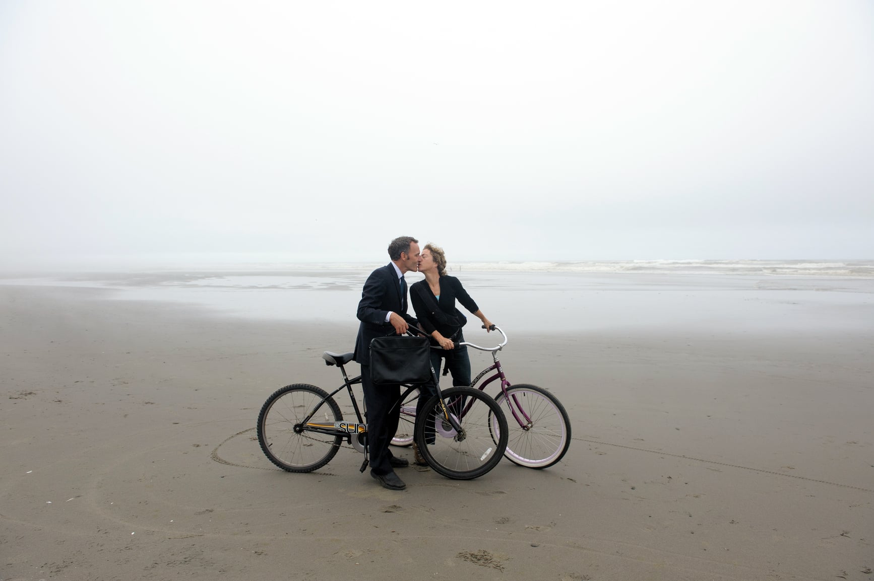 A man and woman sitting on bikes on a misty beach kissing each other.