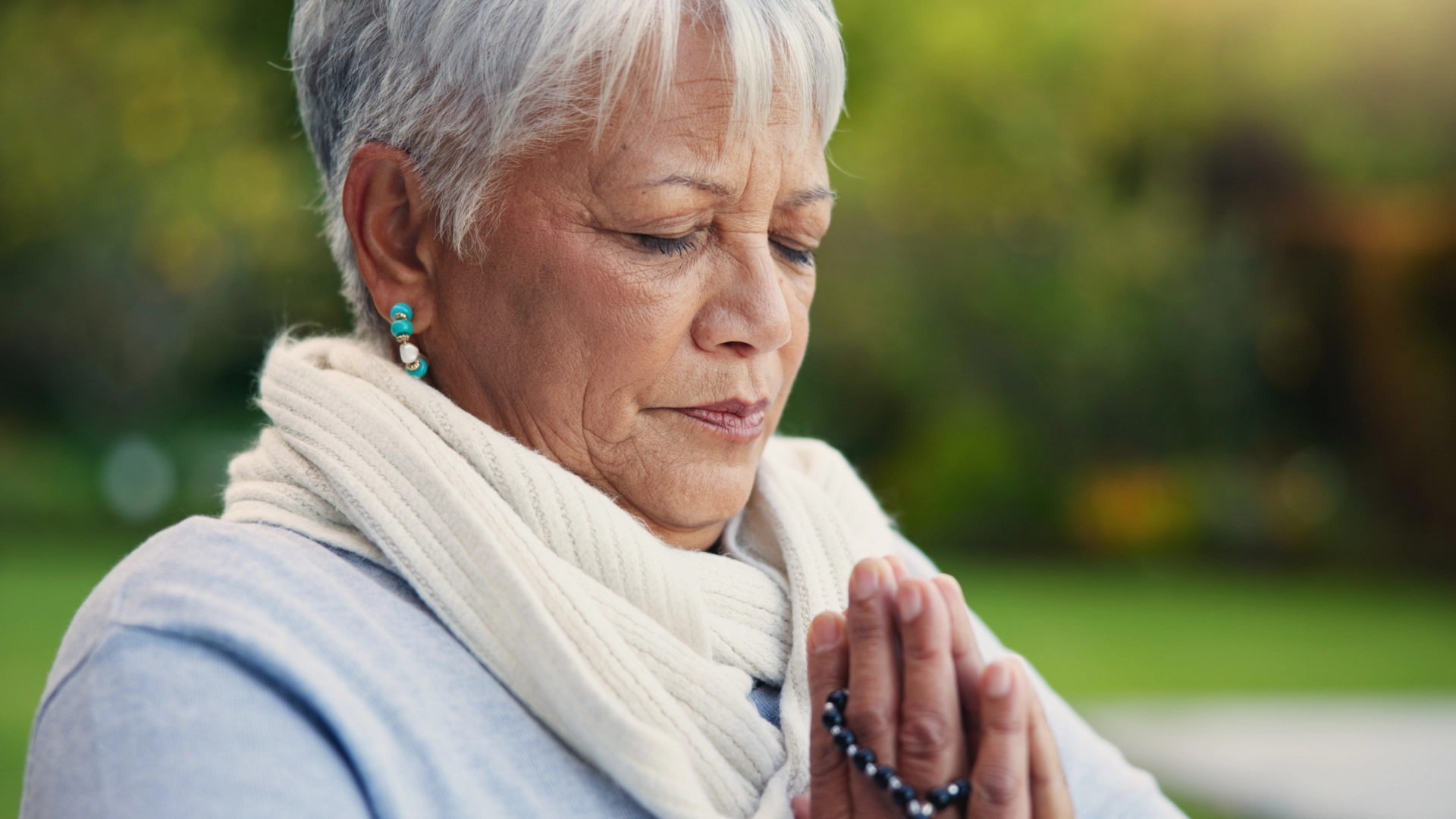 Elderly woman praying with rosary beads outdoors.