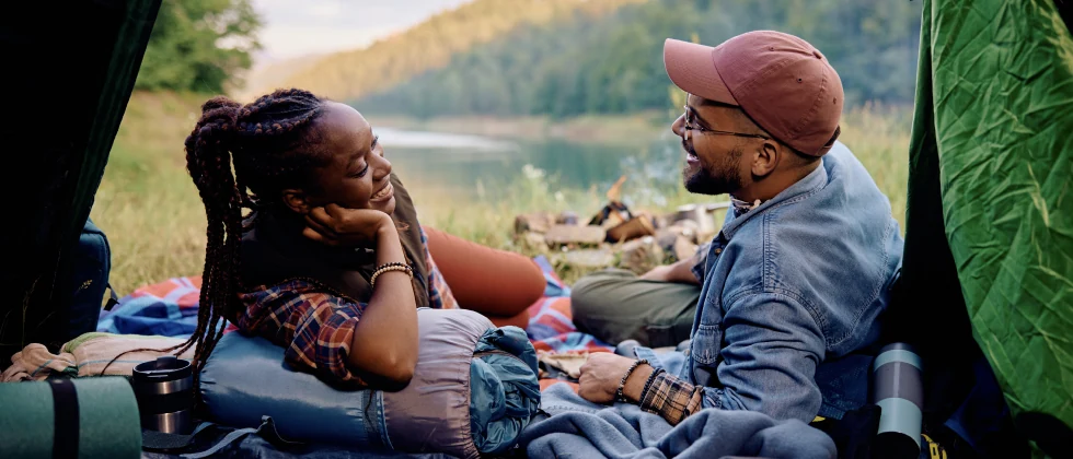 A couple sitting with a tent and picnic blanket in nature, having a conversation.