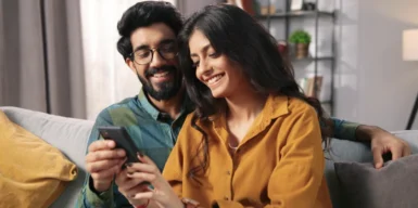 A couple sitting on their couch together an looking at a phone together.