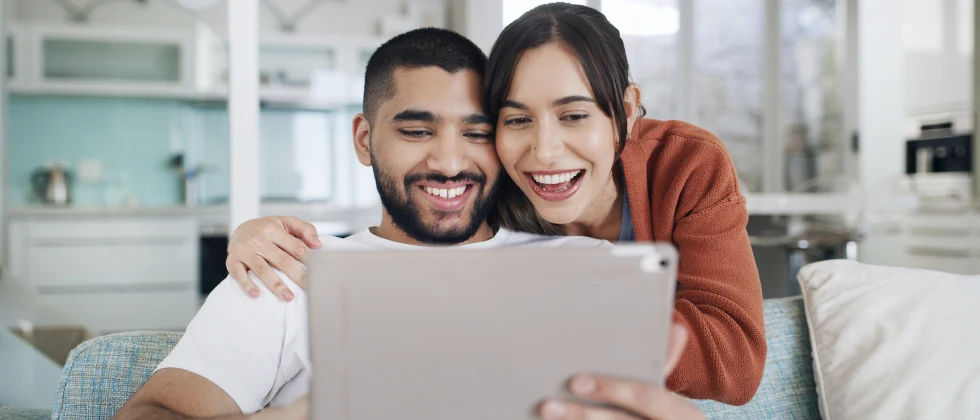 A couple sitting on the couch laughing while looking at a tablet